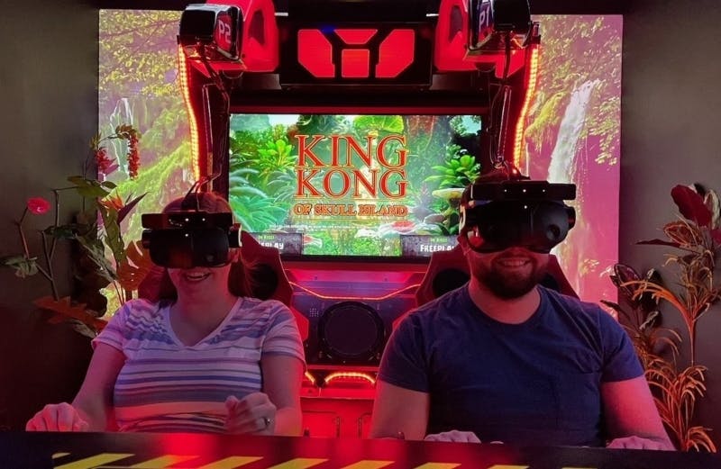 Два человека в VR-гарнитурах сидят в аркадных креслах с красным светом, а на экране позади них отображается «Кинг-Конг: с острова черепа».