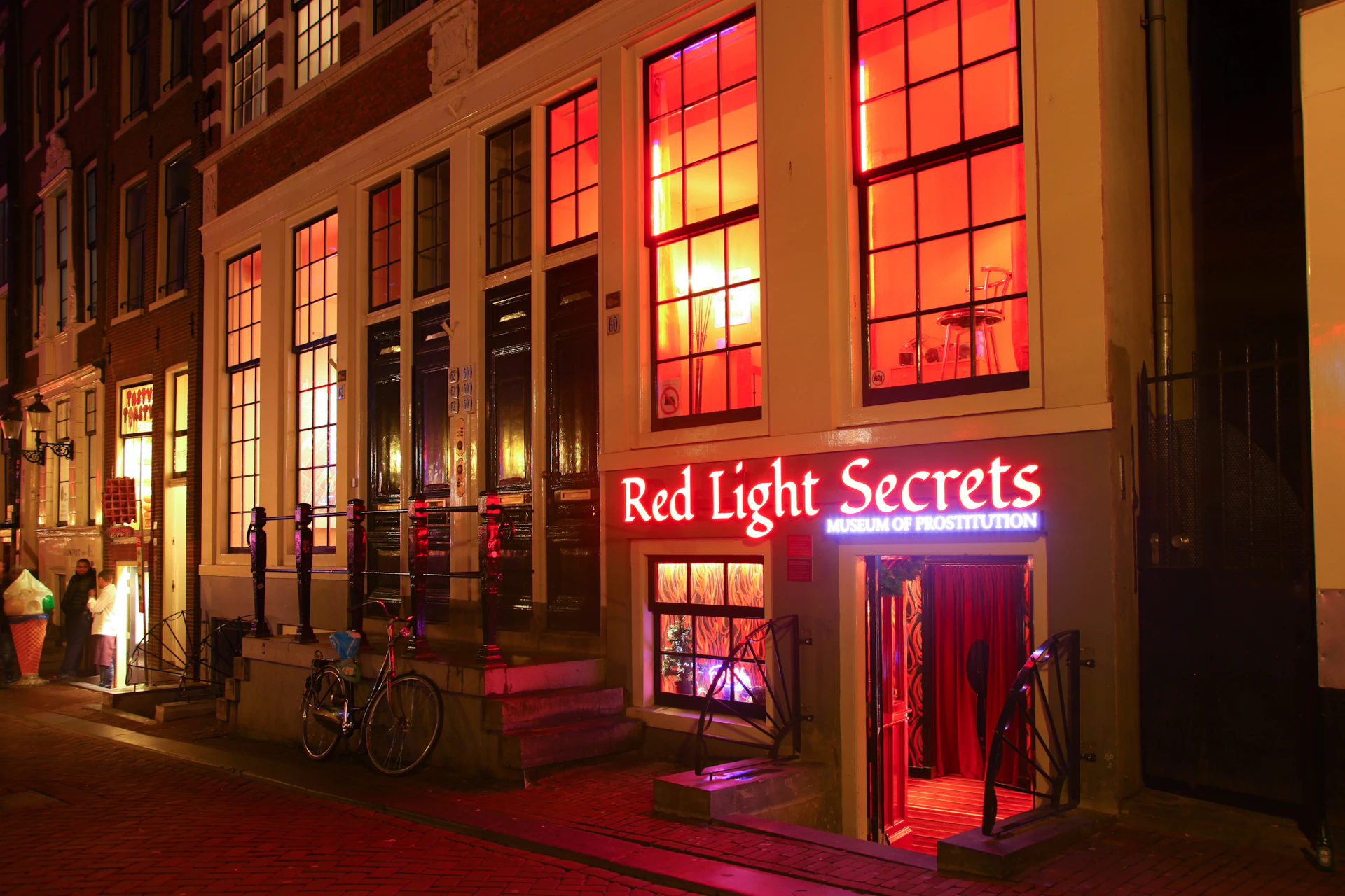 A building with illuminated windows and a neon sign reading "Red Light Secrets Museum of Prostitution" at night.