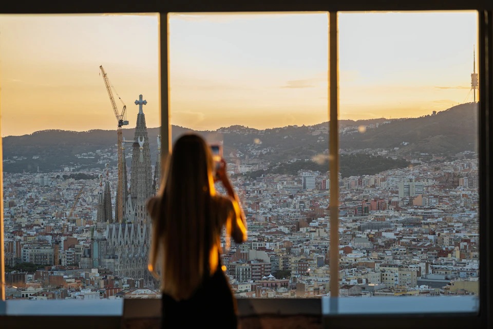 A person with long hair photographs a cityscape at sunset through a large window, with notable buildings in the background.