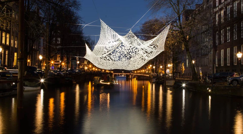 Intricate white crochet art suspended above a canal at night, with reflections of city lights in the water.