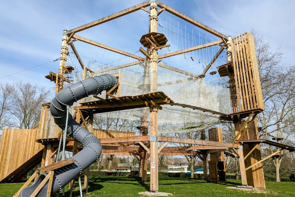 A multi-level outdoor rope course structure with a spiral slide, set on grass against a backdrop of trees and blue sky.