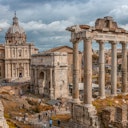 Antiche rovine romane con alte colonne e archi, una chiesa a cupola sullo sfondo e persone che passeggiano nel sito.