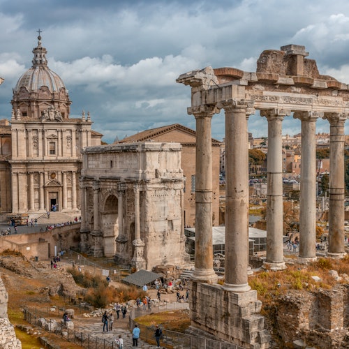 Antiche rovine romane con alte colonne e archi, una chiesa a cupola sullo sfondo e persone che passeggiano nel sito.