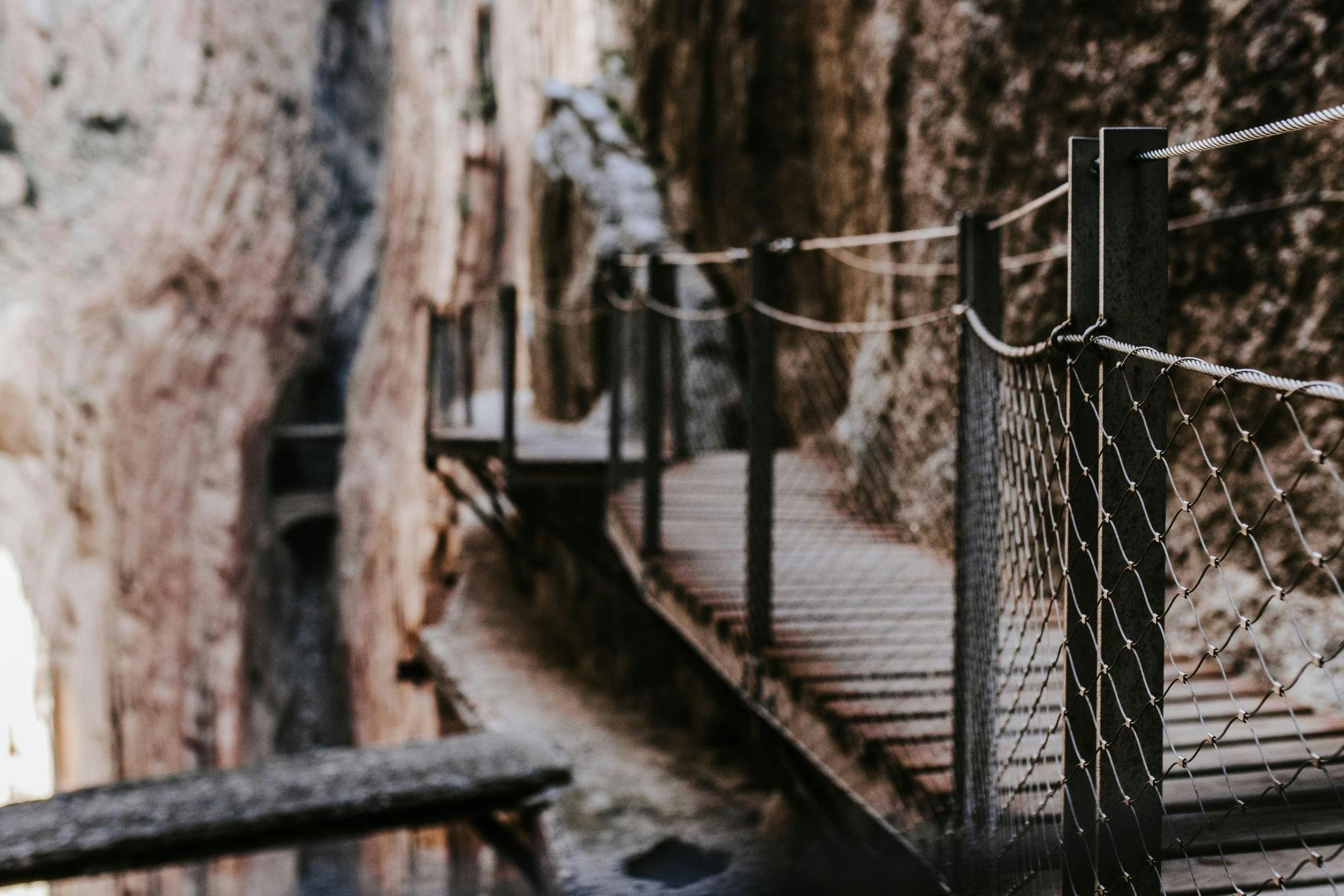 A narrow wooden footbridge with a metal railing spans a rocky canyon, surrounded by rugged cliffs.