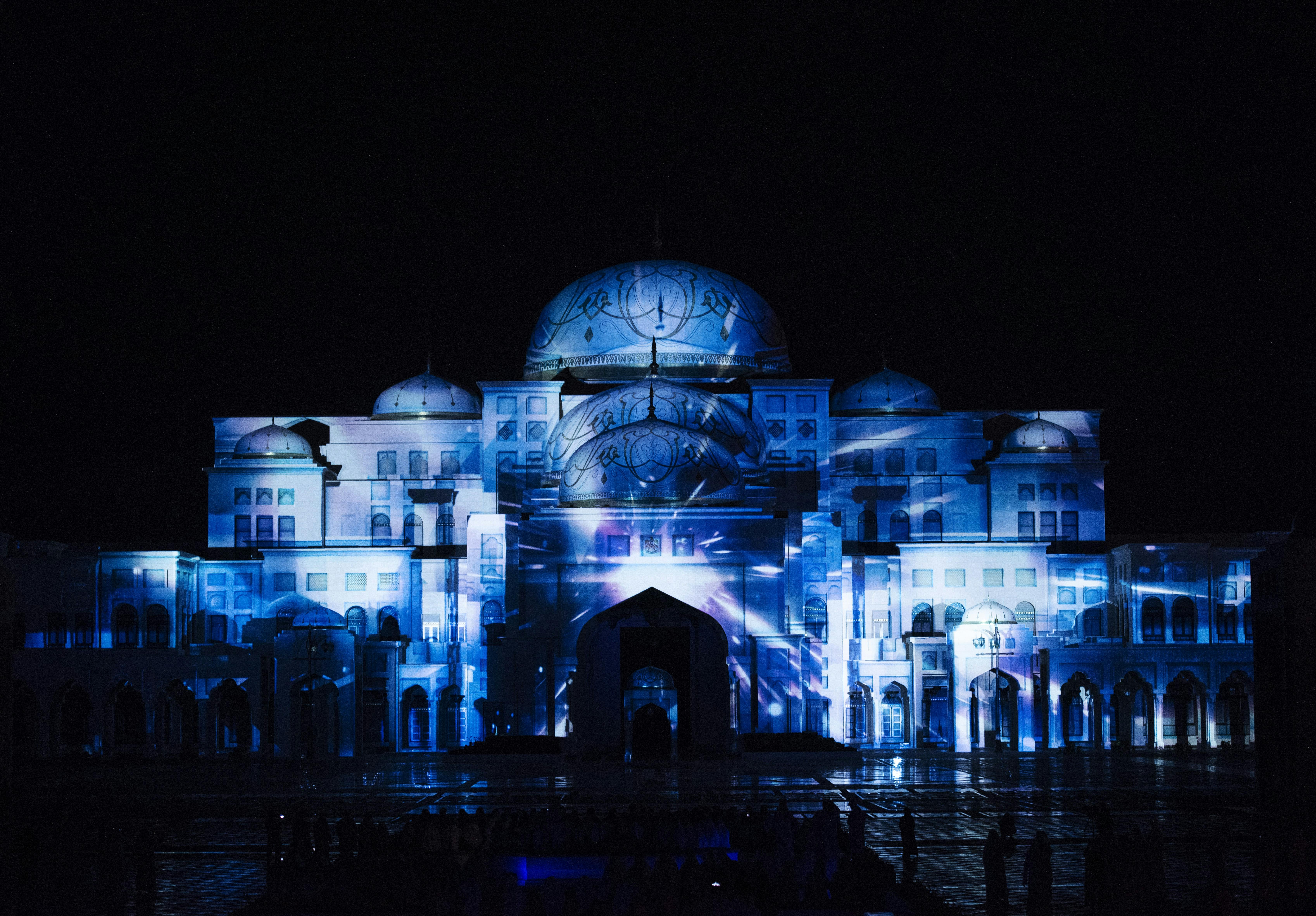 A large illuminated building with domes, arches, and intricate patterns glowing blue against a dark night sky.