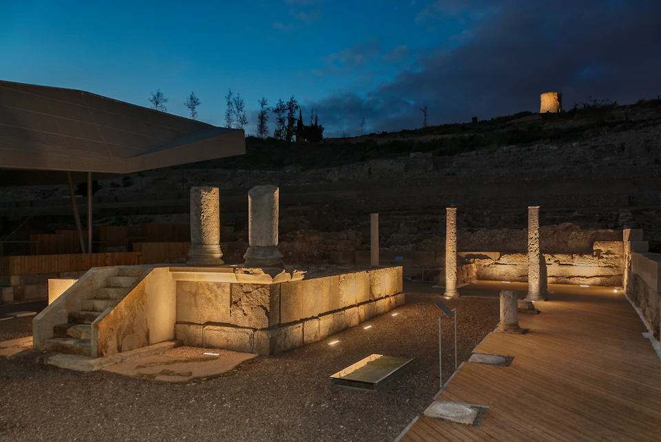 Ancient stone columns and ruins lit up at dusk with a modern concrete building in the background under a darkening sky.