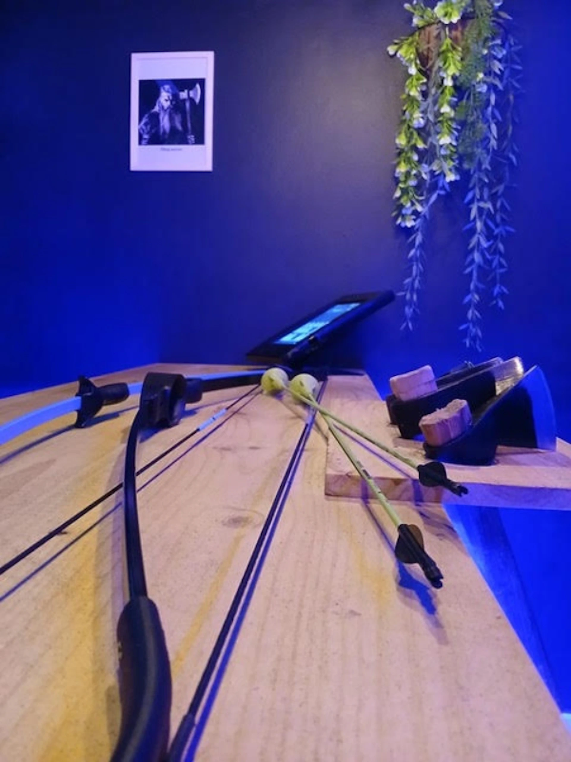 Close-up of a wooden table with two archery bows and a tablet. Wall in background has hanging plants and a framed photo.