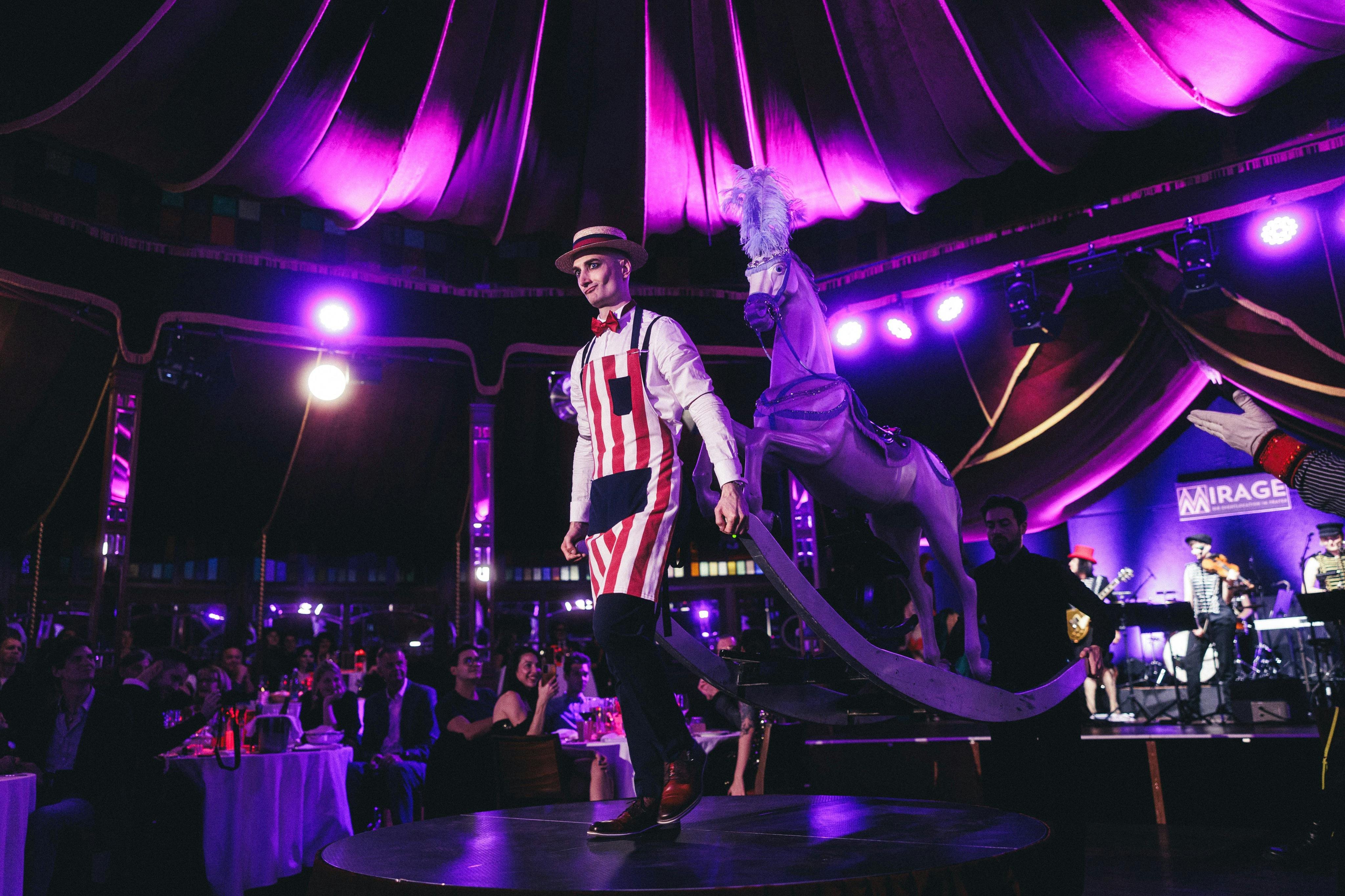 Άντρας με κόκκινη και άσπρη ριγέ στολή και ψάθινο καπέλο που παίζει στη σκηνή δίπλα σε ένα άλογο καρουζέλ, με ένα καθιστό κοινό στο παρασκήνιο.