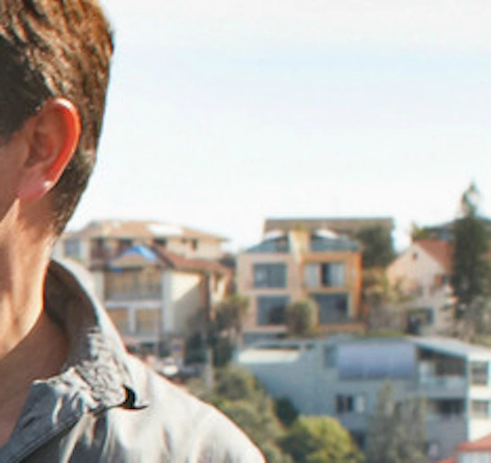 A couple holding hands walks along a coastal path with seaside houses in the background. The man points into the distance.