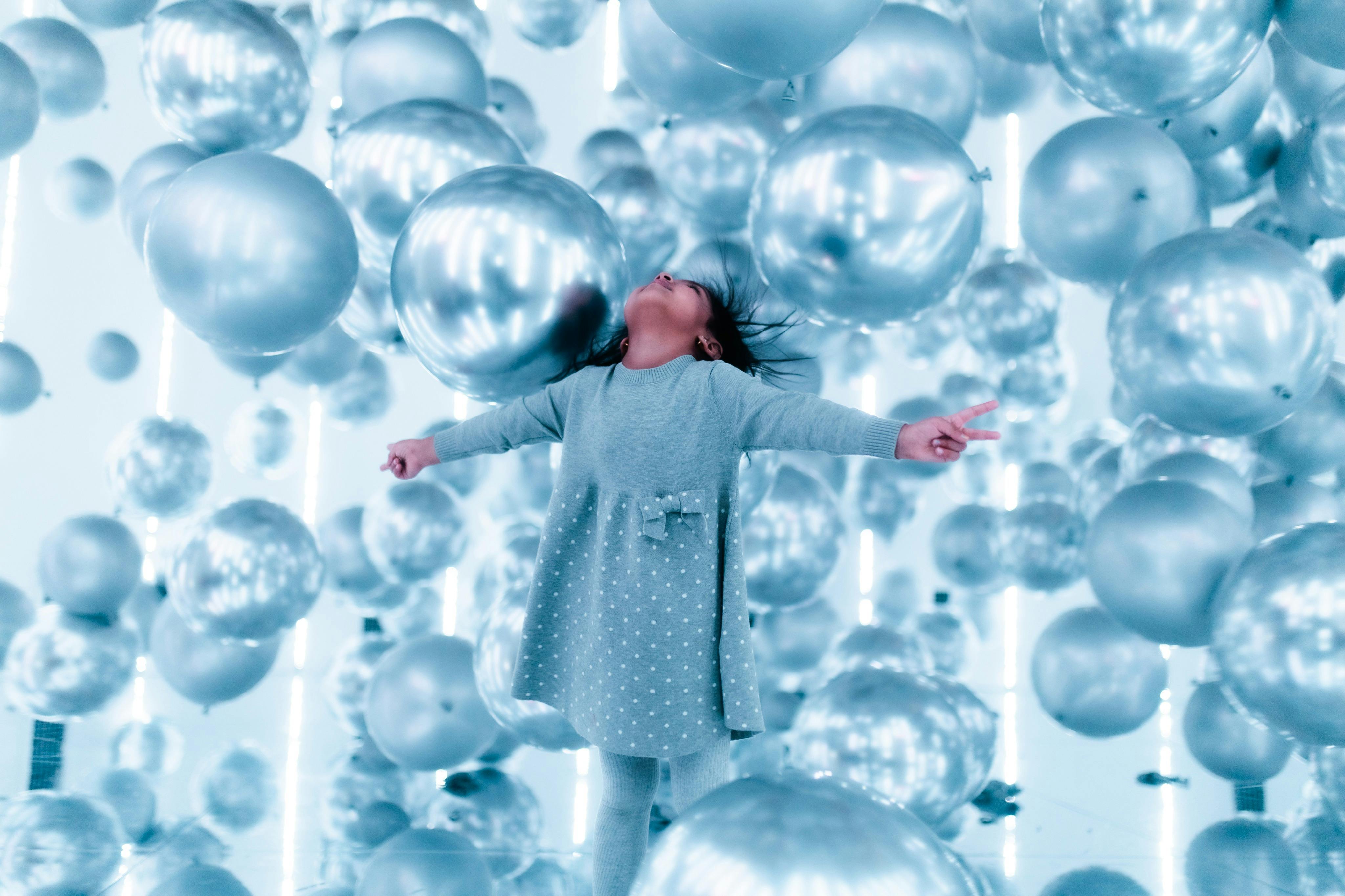 Ребенок в сером наряде радостно позирует среди многочисленных серебряных шариков в ярком, отражающем пространстве.
