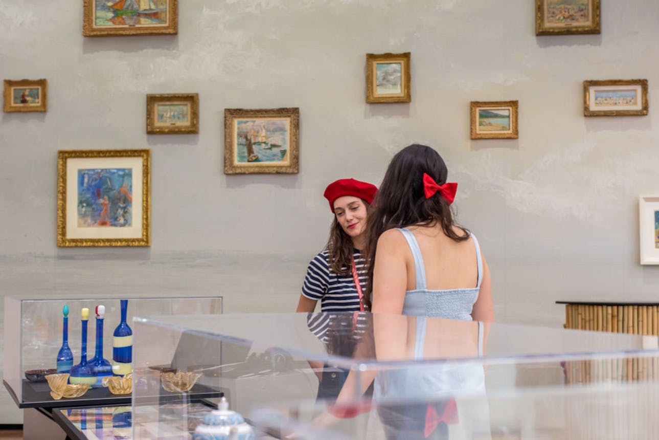 Dvě ženy stojí v umělecké galerii, jedna s červeným baretem a druhá s červenou mašlí. Na zdi za nimi visí zarámované obrazy.