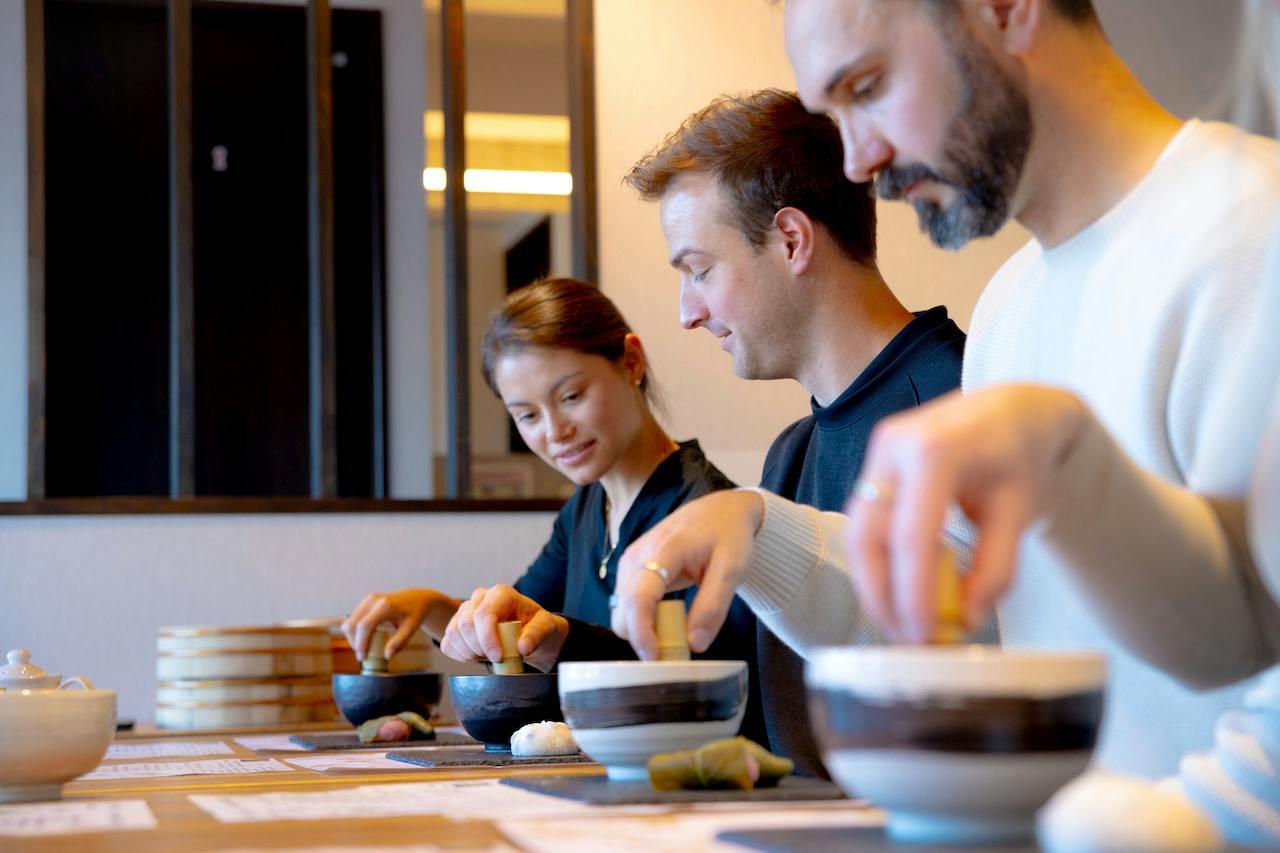 Three people seated at a table preparing food using bowls and ingredients, focusing on their activities.