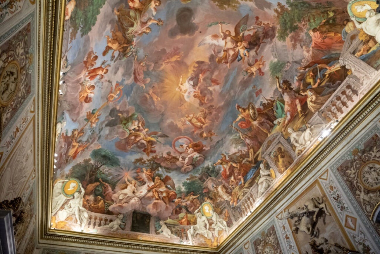 Un soffitto riccamente dipinto con un affresco in stile barocco con figure mitologiche e divine tra nuvole e colori vivaci.