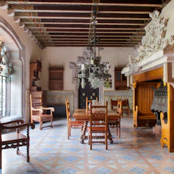 Utsmyckat rum med trästolar och bord, invecklat tak, stor öppen spis och glasmålade välvda fönster.