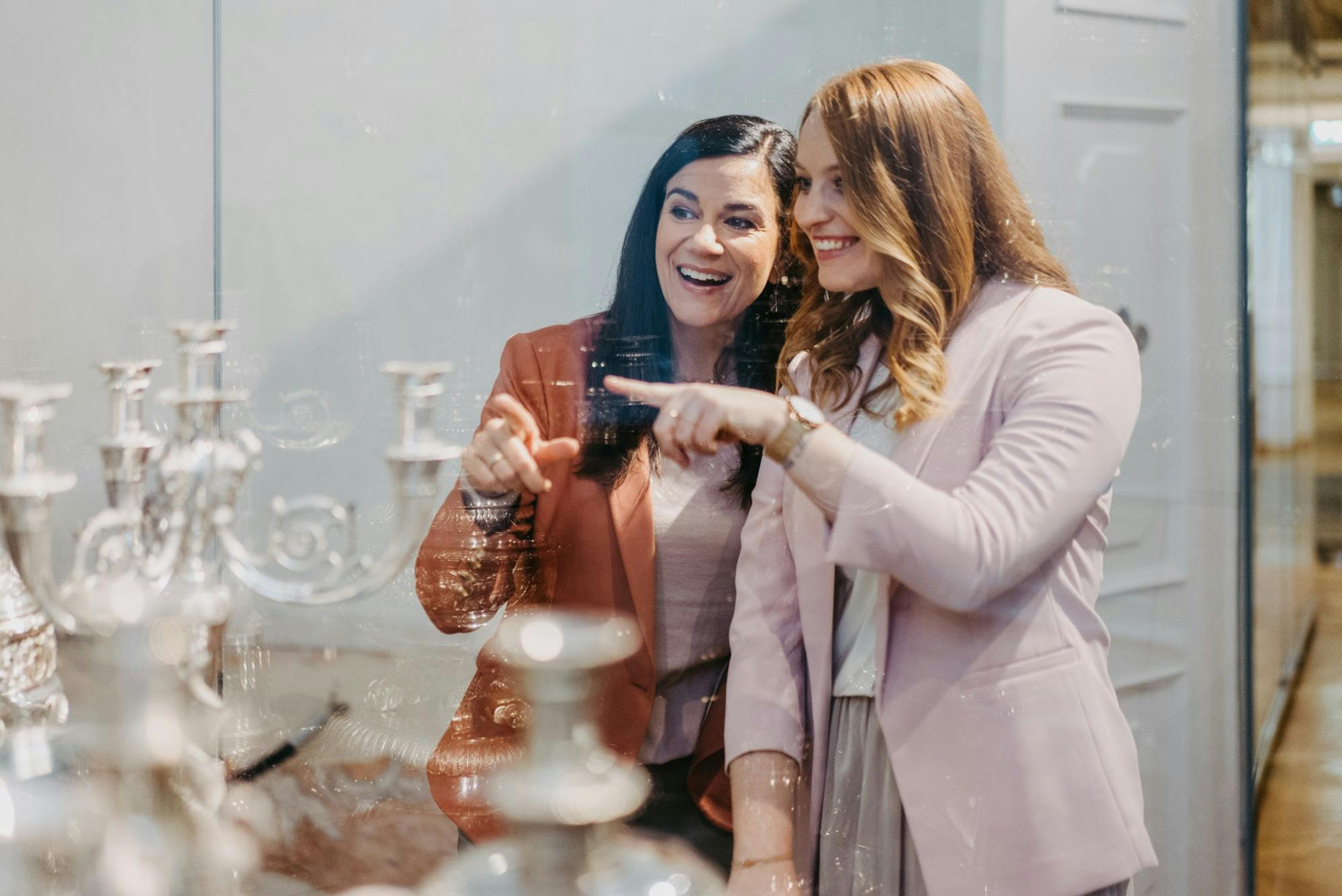 Δύο γυναίκες με σακάκια χαμογελούν και δείχνουν κάτι μπροστά τους, με θολά αντικείμενα στο προσκήνιο.