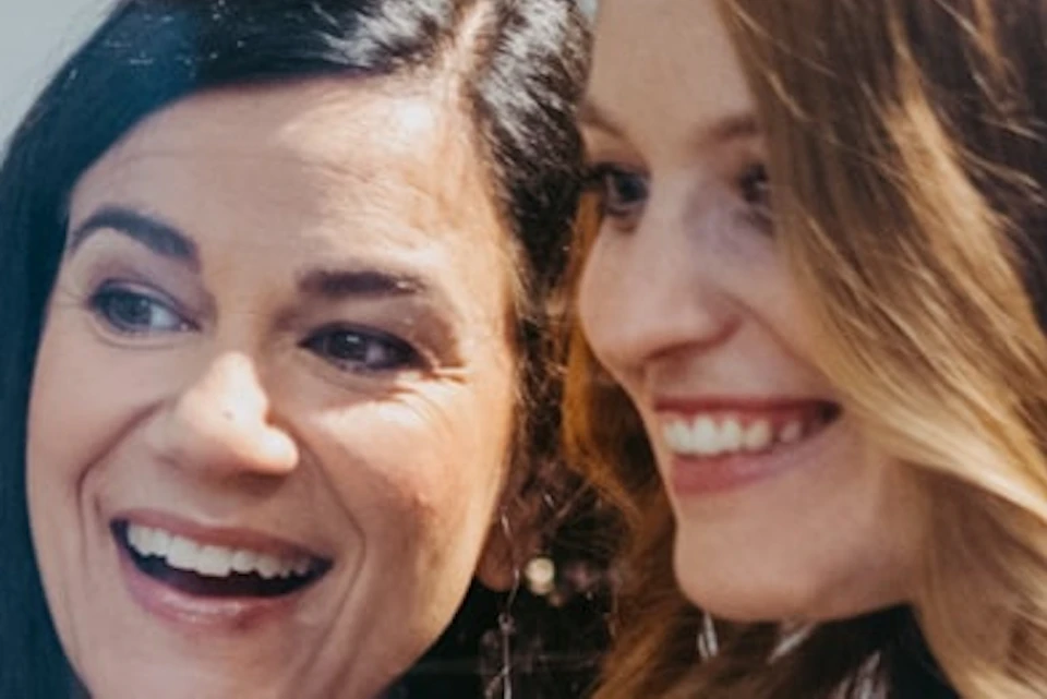 Two women in blazers smiling and pointing at something in front of them, with blurred objects in the foreground.
