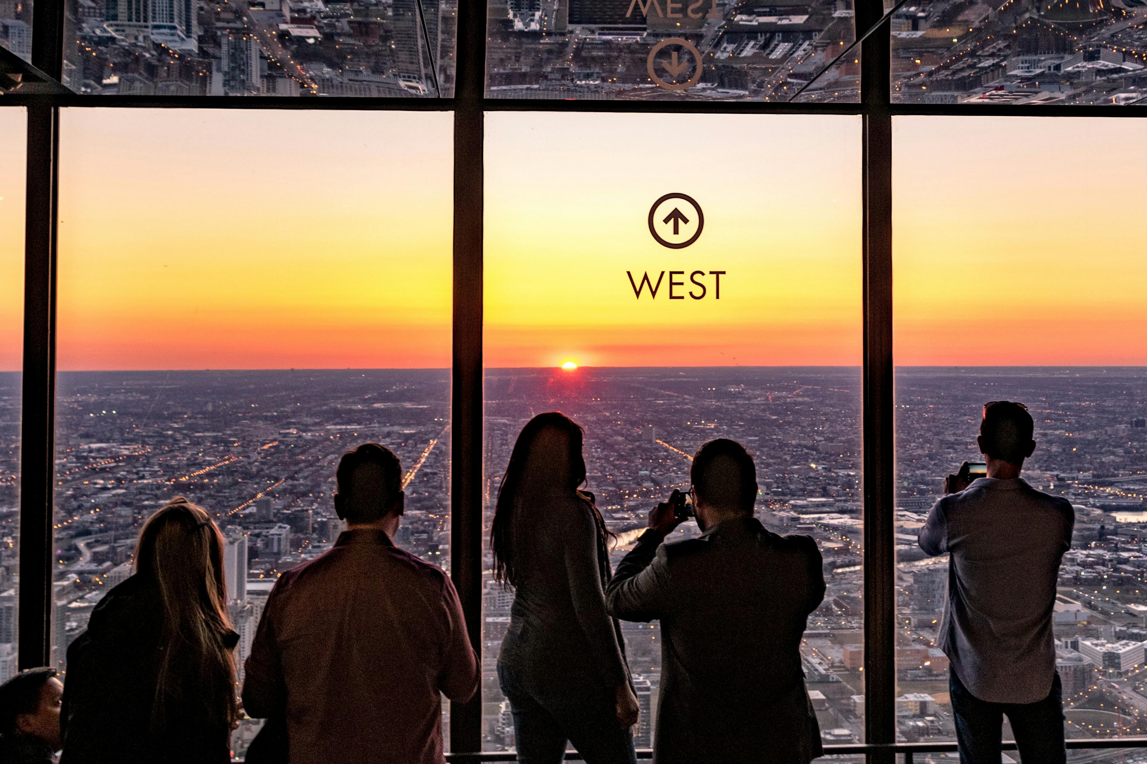 Four people stand inside a high-rise building, watching the sunset over a cityscape through large windows marked "West."