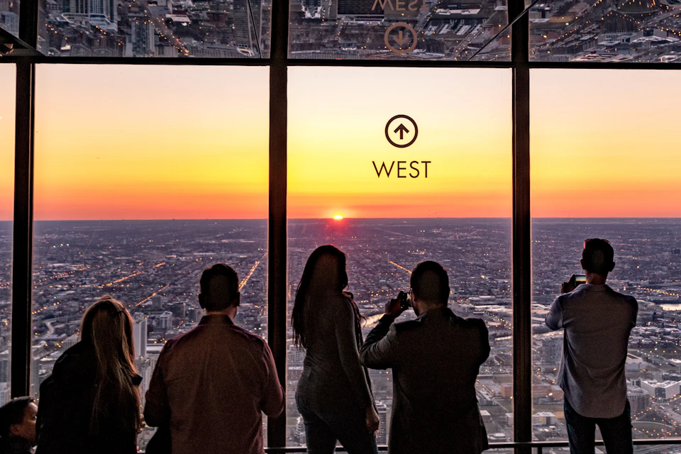 Four people stand inside a high-rise building, watching the sunset over a cityscape through large windows marked "West."
