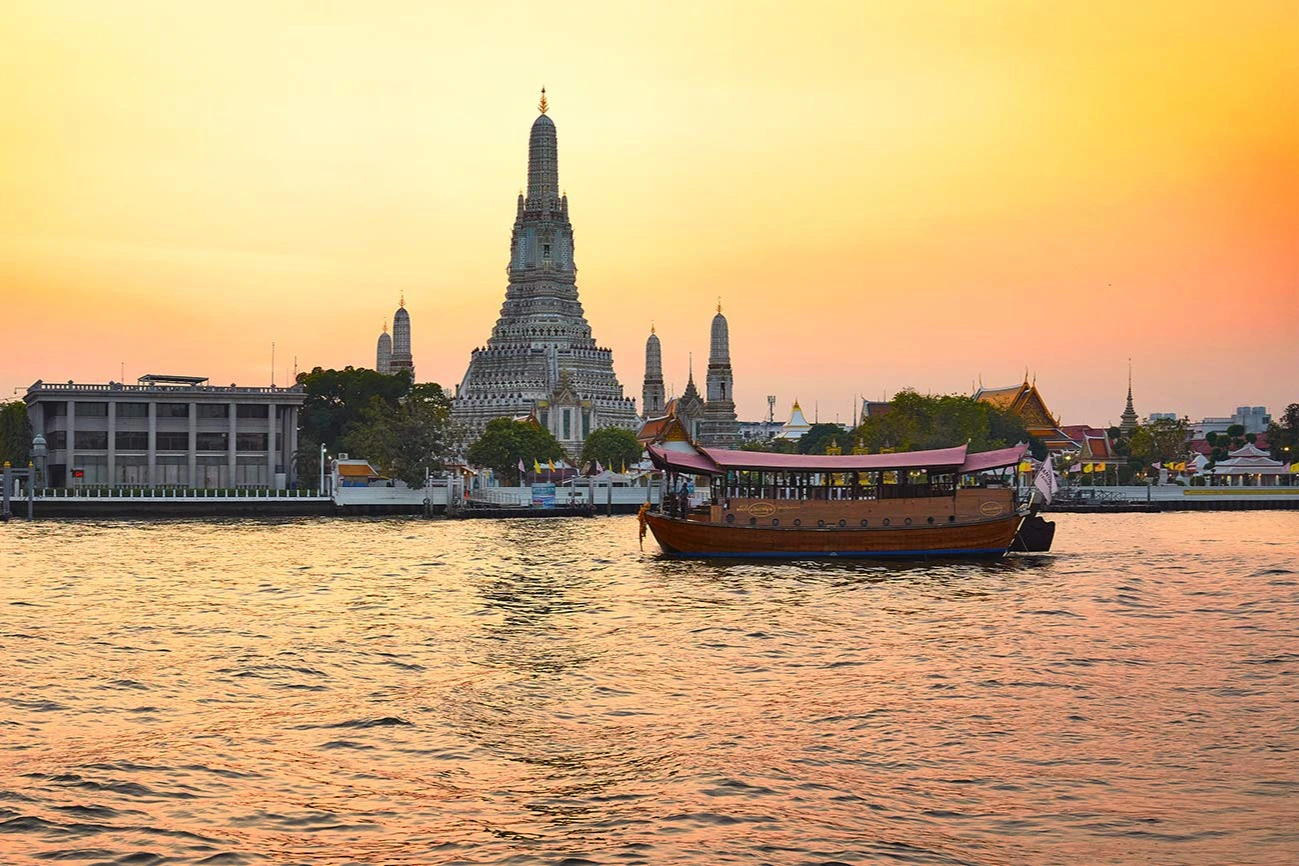 A traditional boat on a river at sunset with a large temple and smaller spires in the background.