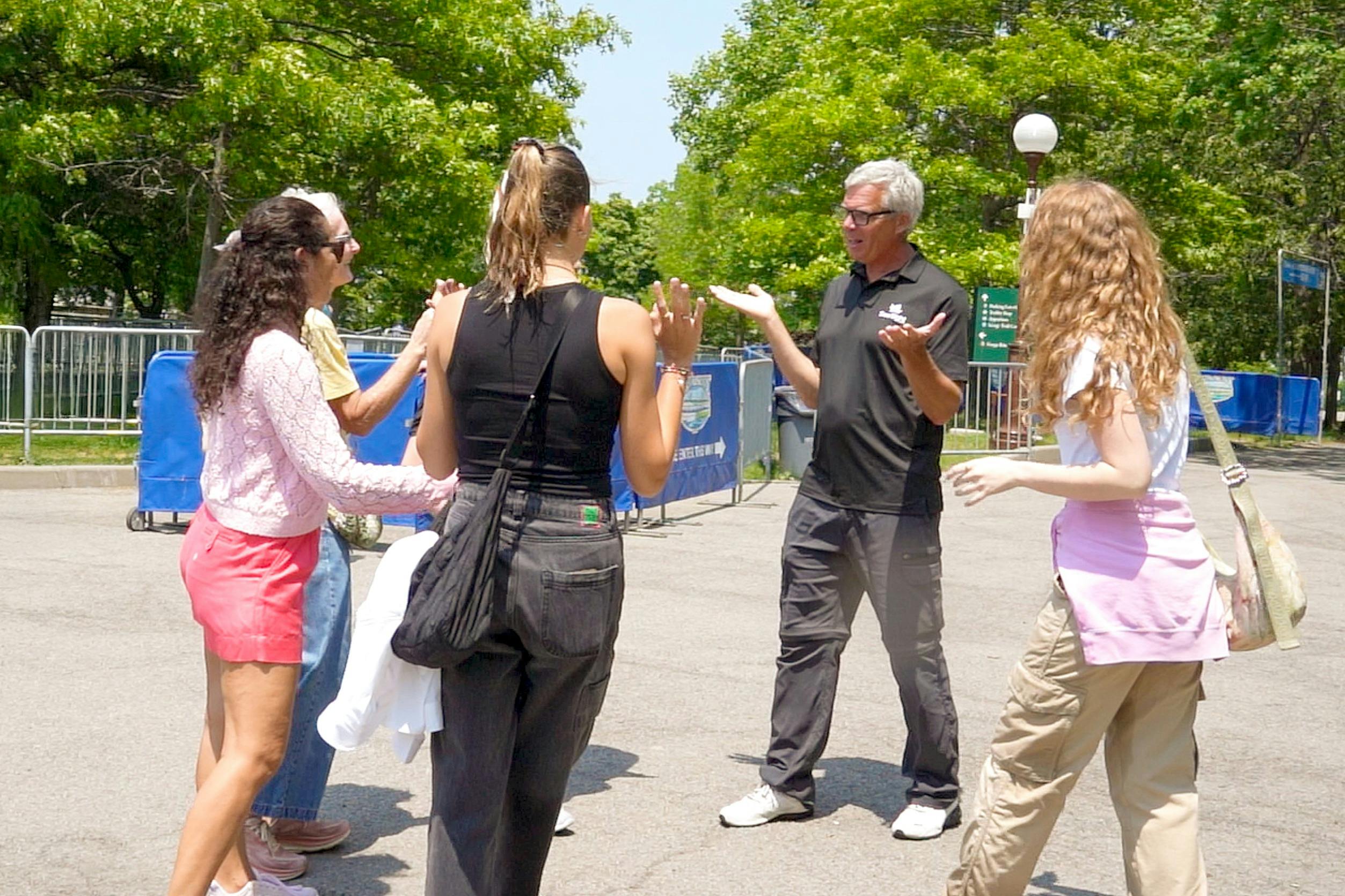 天気のいい日には、男性1人と女性2人の3人が屋外で会話をしています。背景には木々と街灯柱があります。