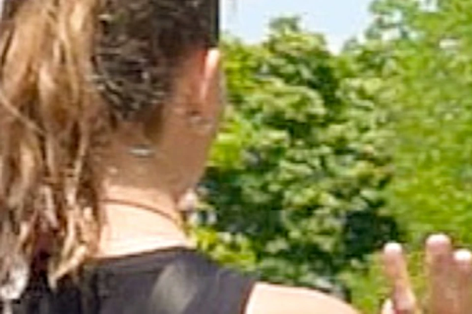 Three people, one man and two women, converse outdoors on a sunny day with trees and a lamppost in the background.