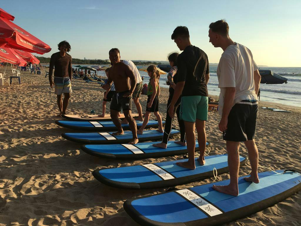 Grupa osób stojących na niebieskich deskach surfingowych leżała na piaszczystej plaży, pod częściowo zachmurzonym niebem podczas zachodu słońca.