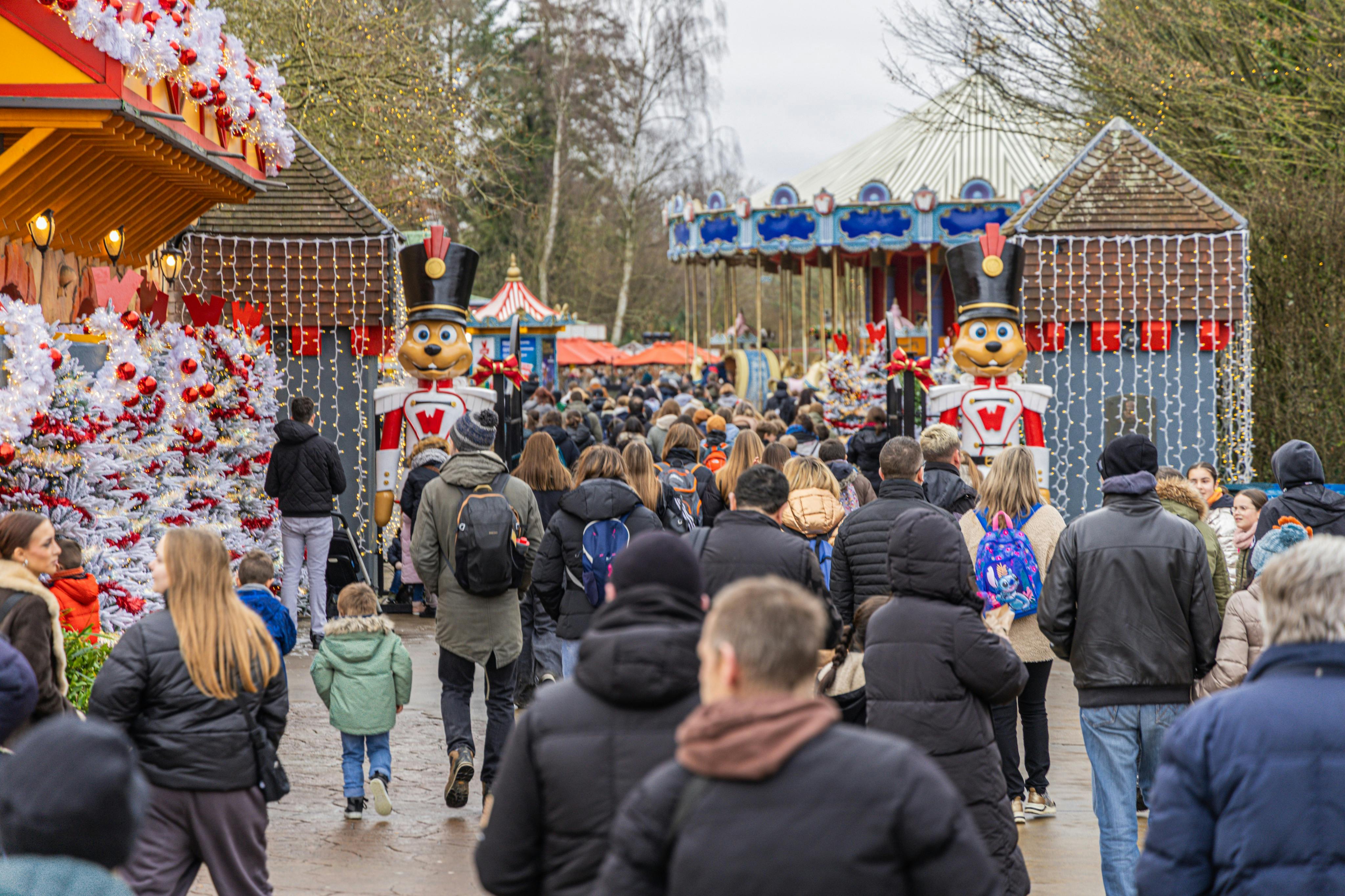 Dav lidí kráčí směrem k slavnostnímu zábavnímu parku s dekoracemi, včetně velké figurky vojáčka a kolotoče.