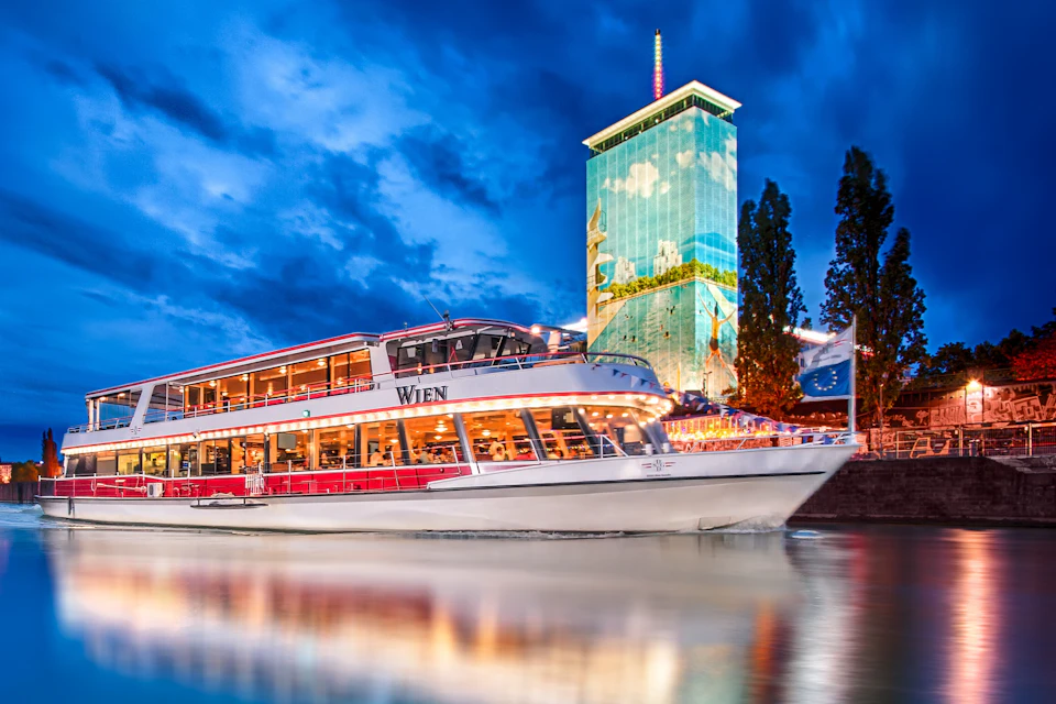 A brightly lit riverboat named "Wien" is docked with a tall, illuminated building in the background during dusk.