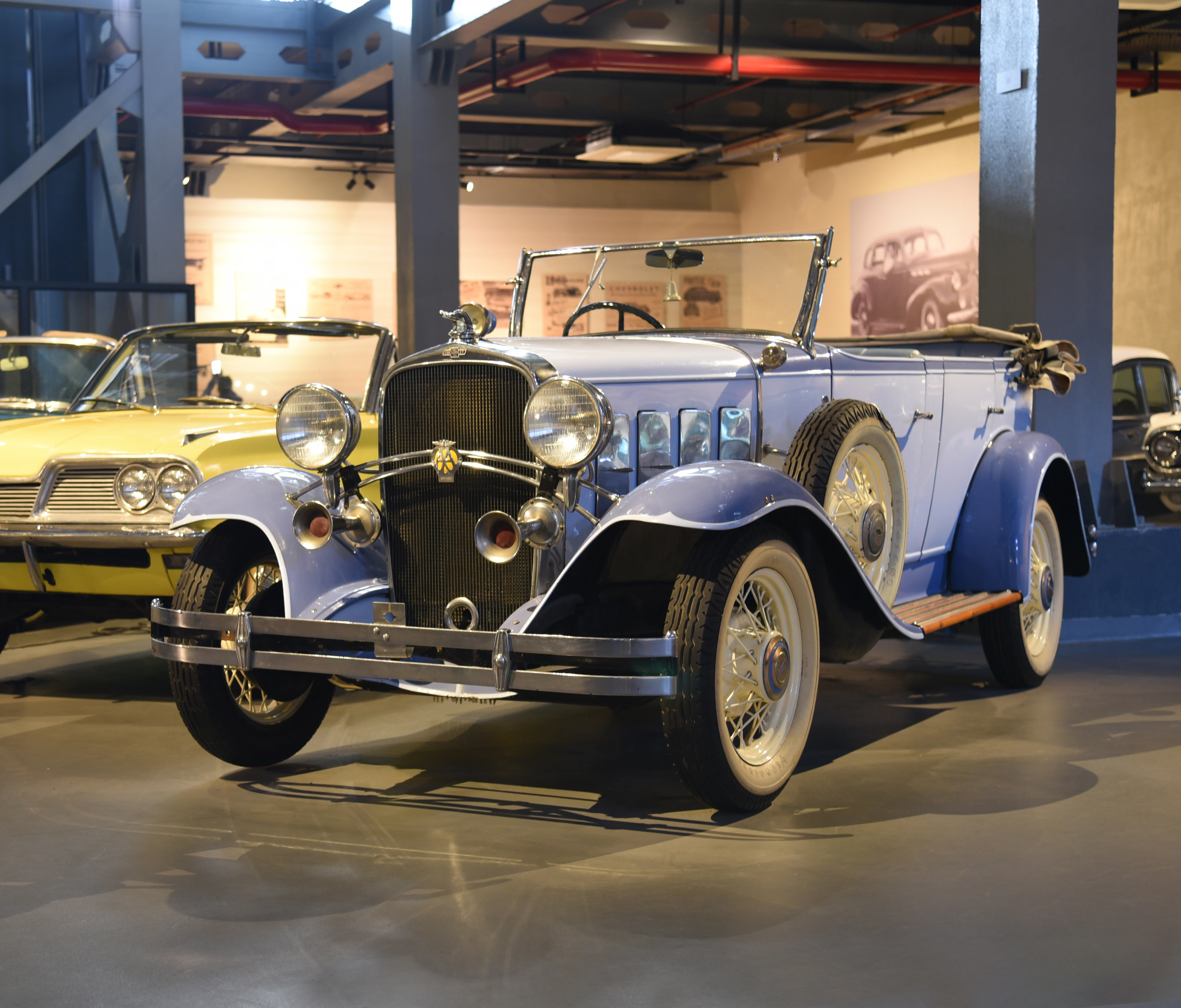 A vintage blue convertible car with large headlights and wire wheels is displayed in an indoor exhibition space.