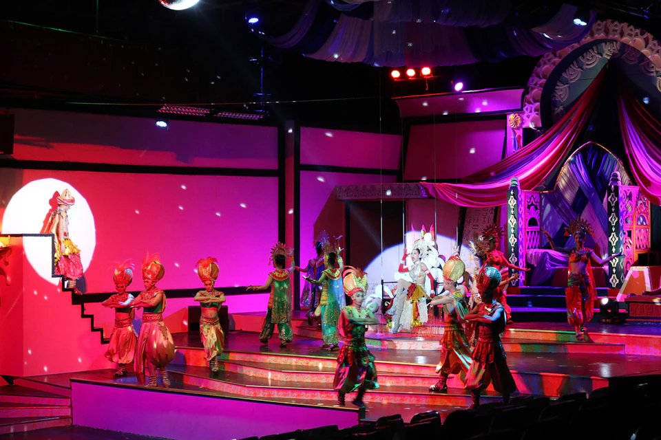 A vibrant theater stage with performers in colorful costumes dancing under bright lights and decorative backdrops.