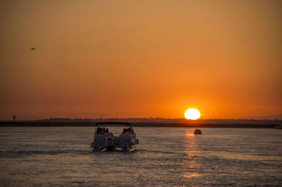 A boat cruising on the water towards a vibrant sunset, with the sun low on the horizon and the sky glowing orange