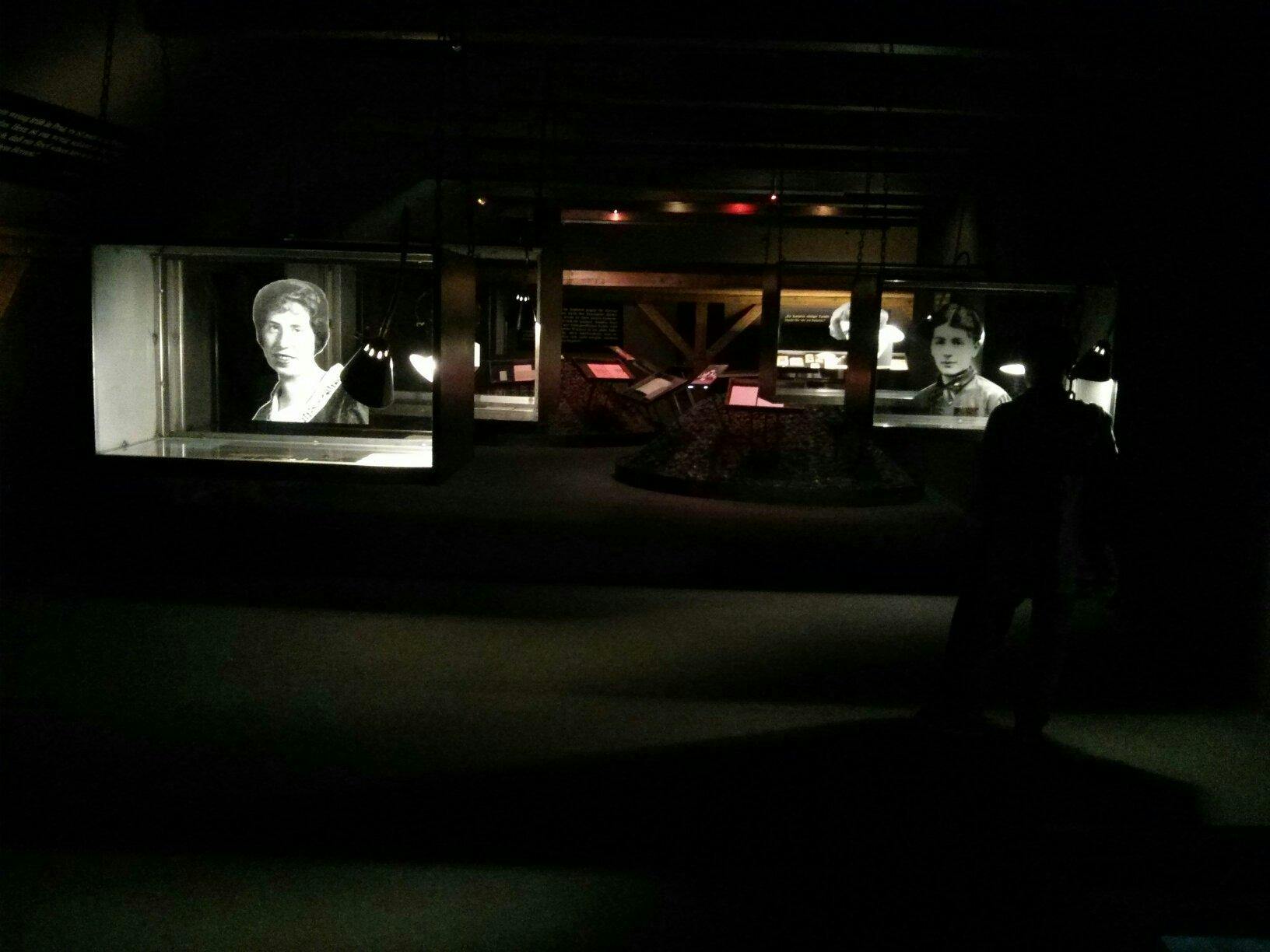 Exposition muséale faiblement éclairée avec des photographies historiques en noir et blanc et divers panneaux d'information sur des présentoirs.