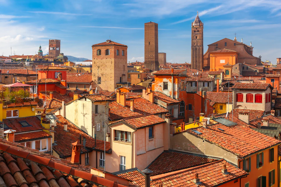 A cityscape of Bologna, Italy, featuring red-roofed buildings, towers, and clear blue skies in the background.