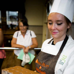A chef in a white hat and apron holding a tray, while a woman in the background watches, holding a green folder.