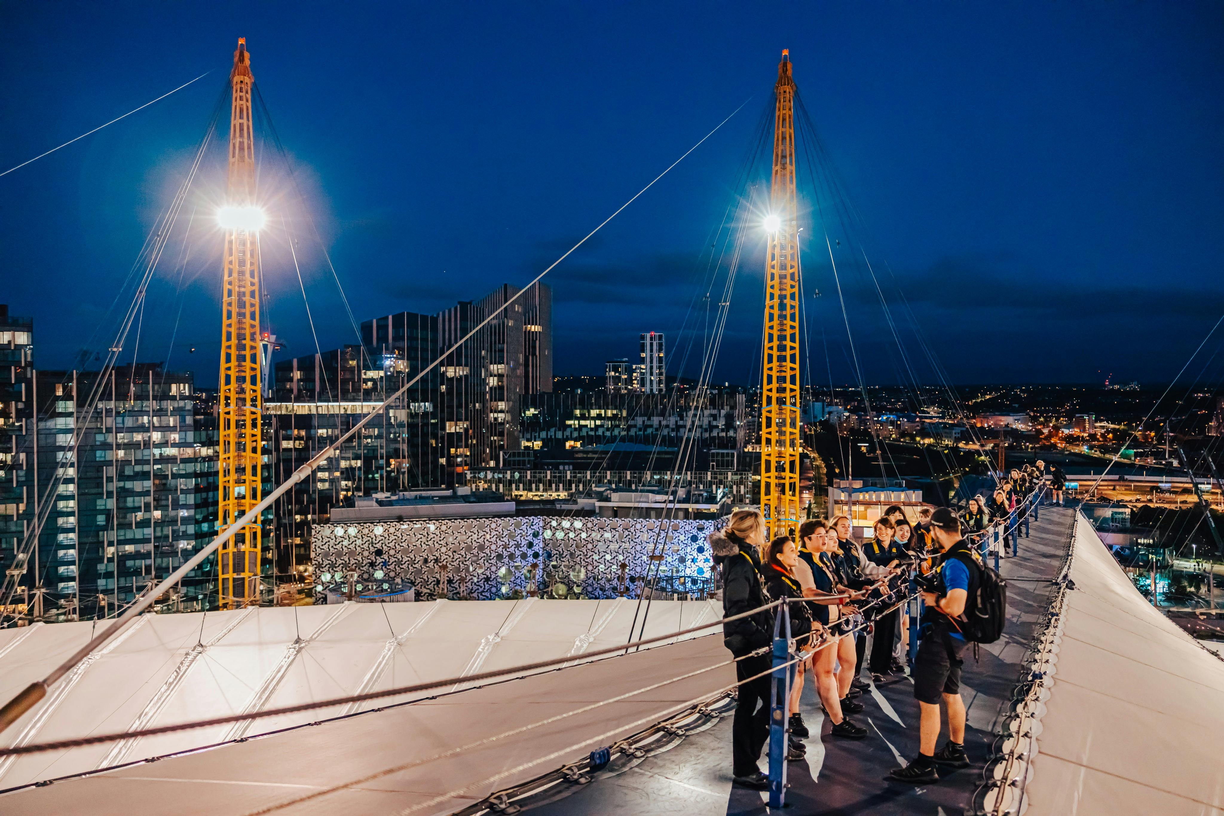 Skupina lidí stojí v noci na zavěšeném chodníku a shlíží na panoráma města s budovami a osvětlenými strukturami.