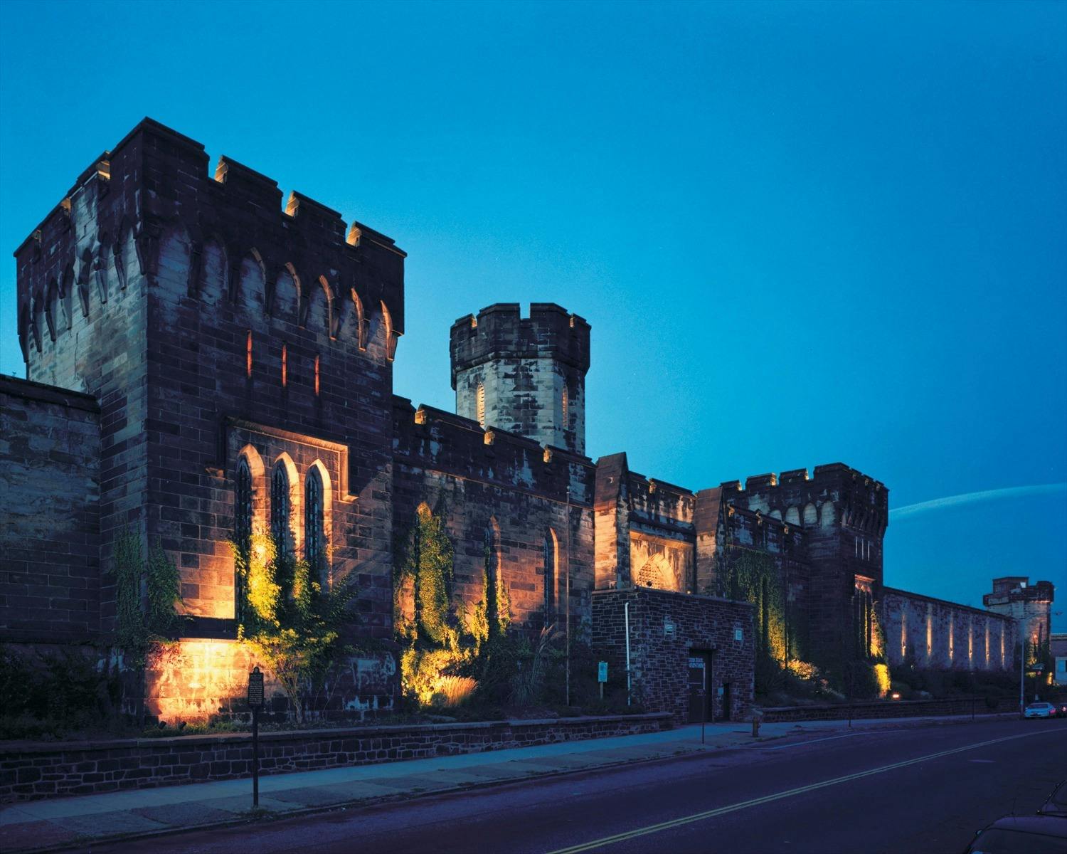 Un gran castillo de piedra iluminado con múltiples torreones y torres al anochecer, visto desde una calle cercana.