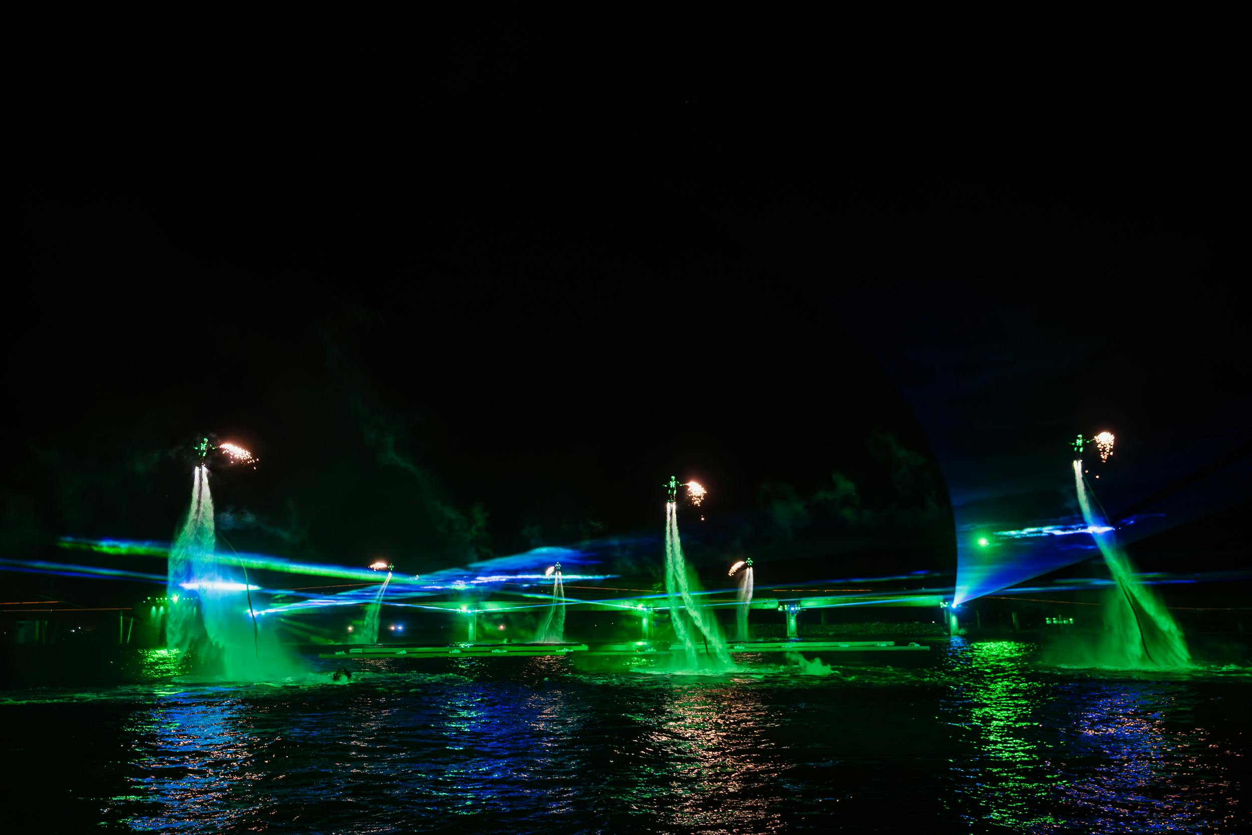 Nighttime water show with green lights, fountains, and performers on jetpacks emitting fire over a body of water.