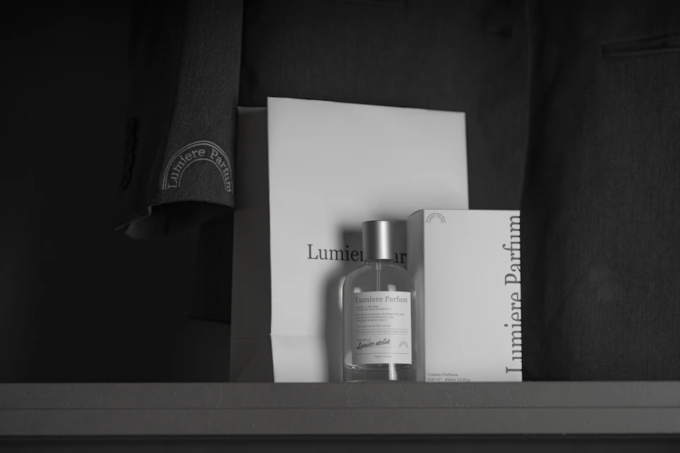 A bottle and box of Lumiere Parfum placed on a shelf, with a branded bag and jacket in the background.