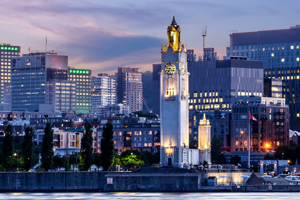 A scenic view of a city at dusk with a lit clock tower near the waterfront and modern buildings in the background.