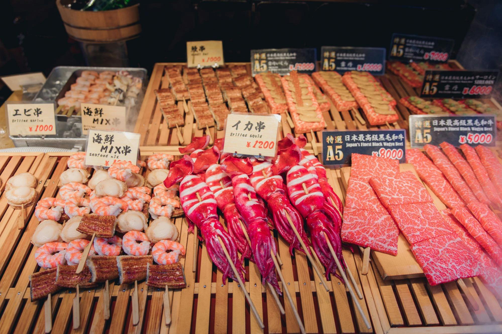A display of skewered seafood, including squid and shrimp, with price signs in Japanese, on a wooden rack at a market.