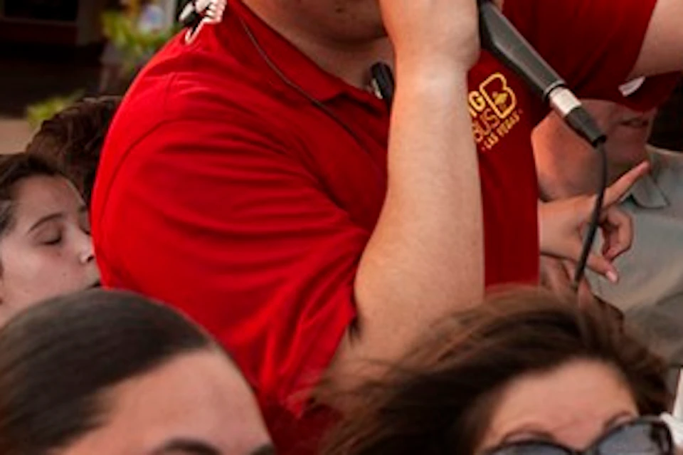 A tour guide in a red shirt speaks into a microphone while pointing. Several smiling passengers are on an open-top bus.