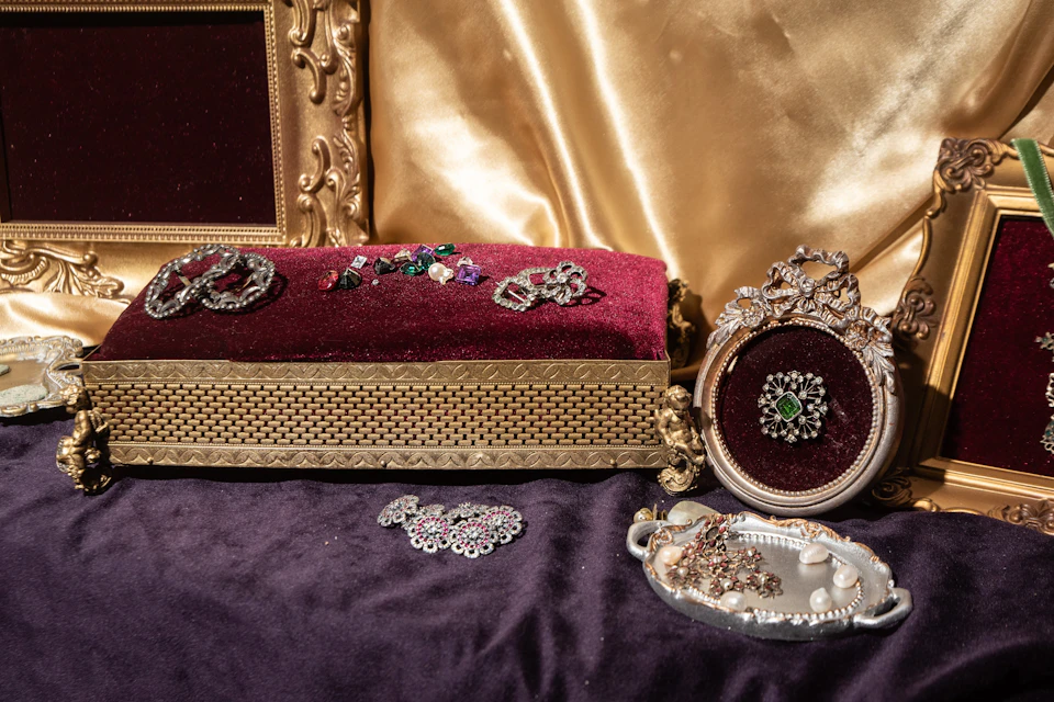 Ornate jewelry including brooches and necklaces displayed on a red velvet jewelry box and small tray, set against a gold satin backdrop.