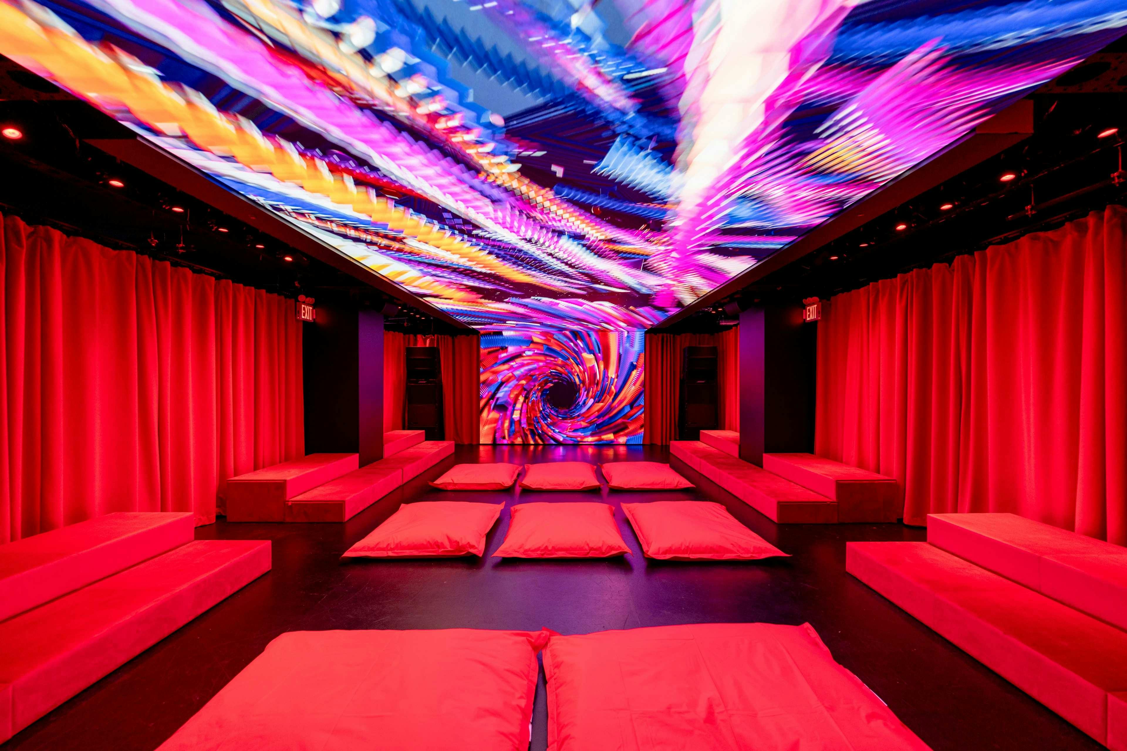 A dimly lit room with red cushions on the floor, red curtains on the sides, and a vibrant, swirling light display on the ceiling and wall.