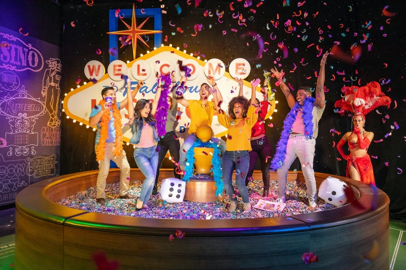 A group of people celebrating with confetti and feather boas in front of a bright "Welcome to Fabulous" sign; performers in colorful costumes.