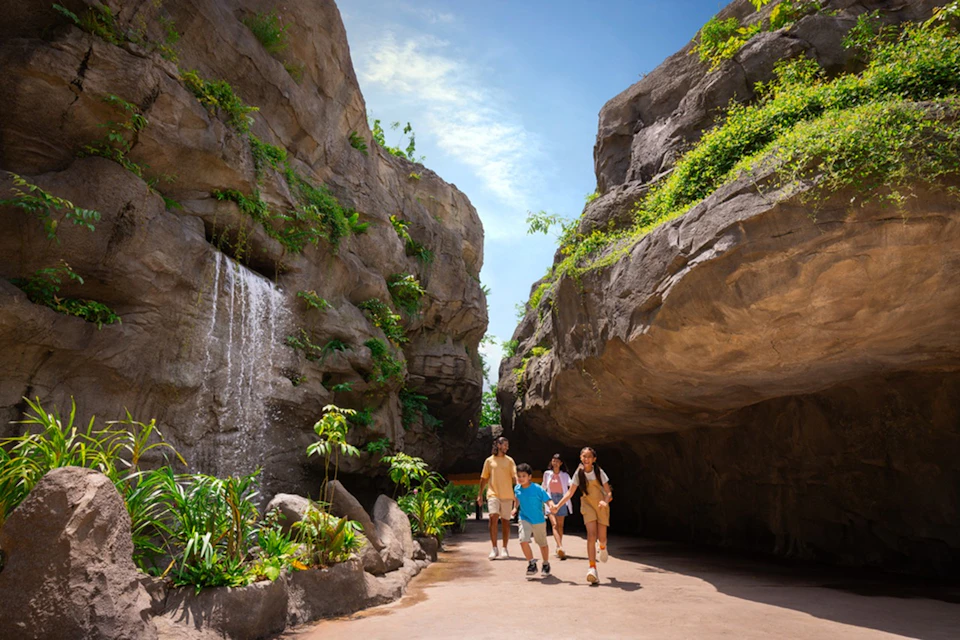 Family walking through a narrow natural rock canyon with greenery and a small waterfall, under a bright blue sky.