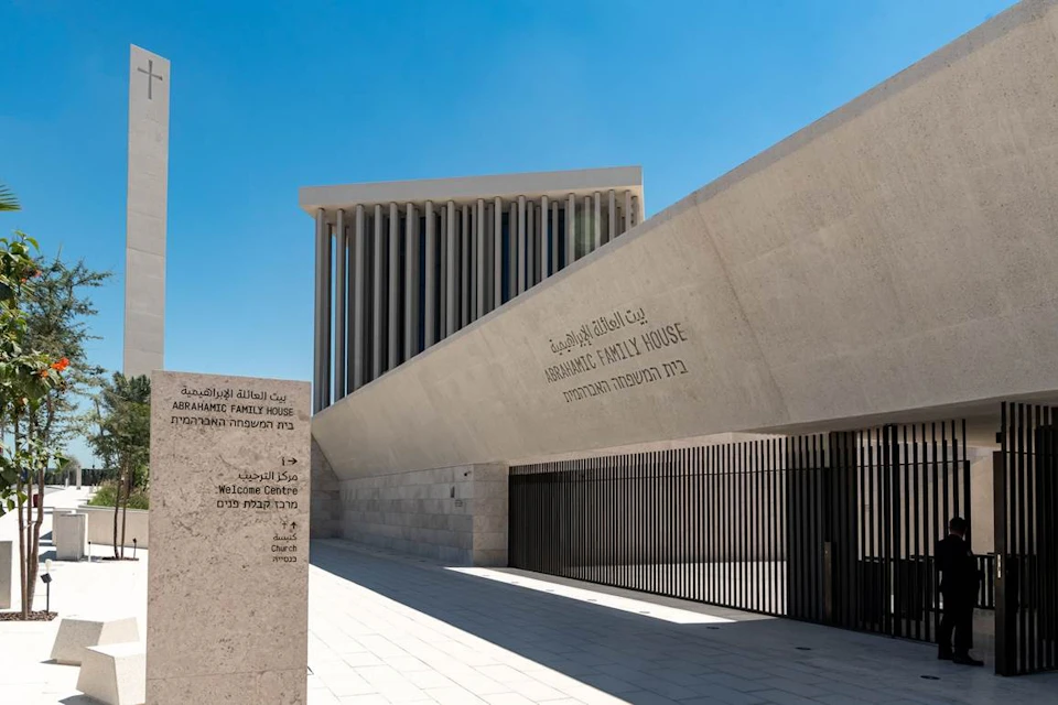 Modern building with the text "Abrahamic Family House" inscribed on an angled wall. Vertical columns and a blue sky are visible.