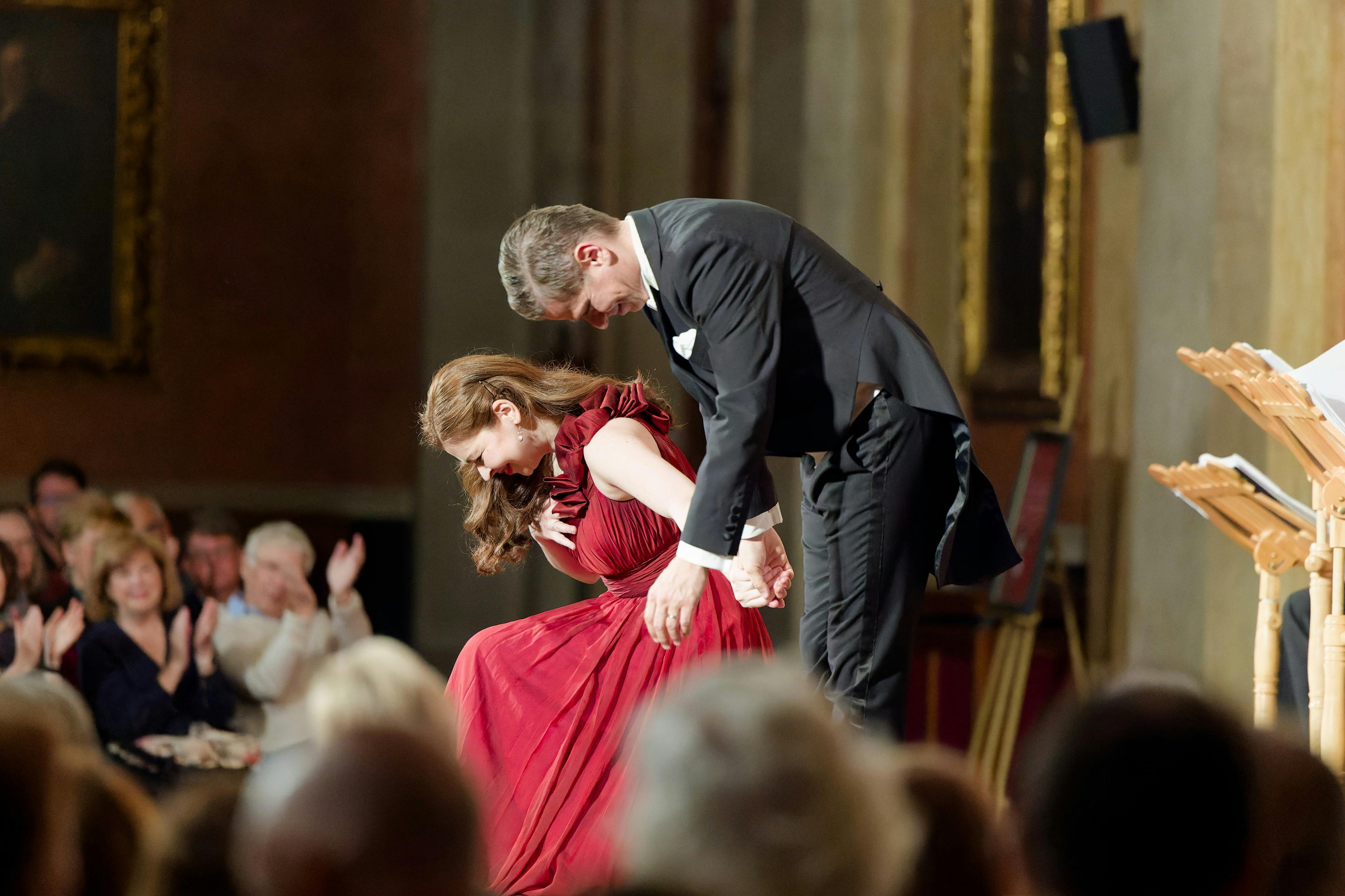 To personer i formelt påklædning bukker på scenen, mens et publikum klapper i et rigt dekoreret interiør.