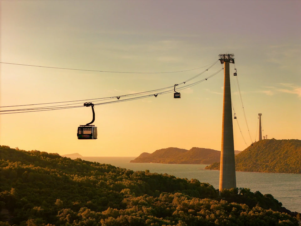 Cable cars suspended over a forested area with water and distant hills at sunset.