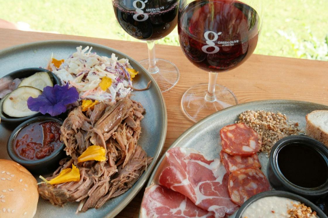 Plates of pulled meat, charcuterie, and salad next to two glasses of red wine on a wooden table in an outdoor setting.