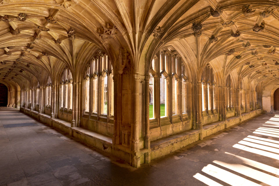 A stone corridor with pointed arches and windows, featuring intricate vaulted ceilings and sunlight casting patterns.
